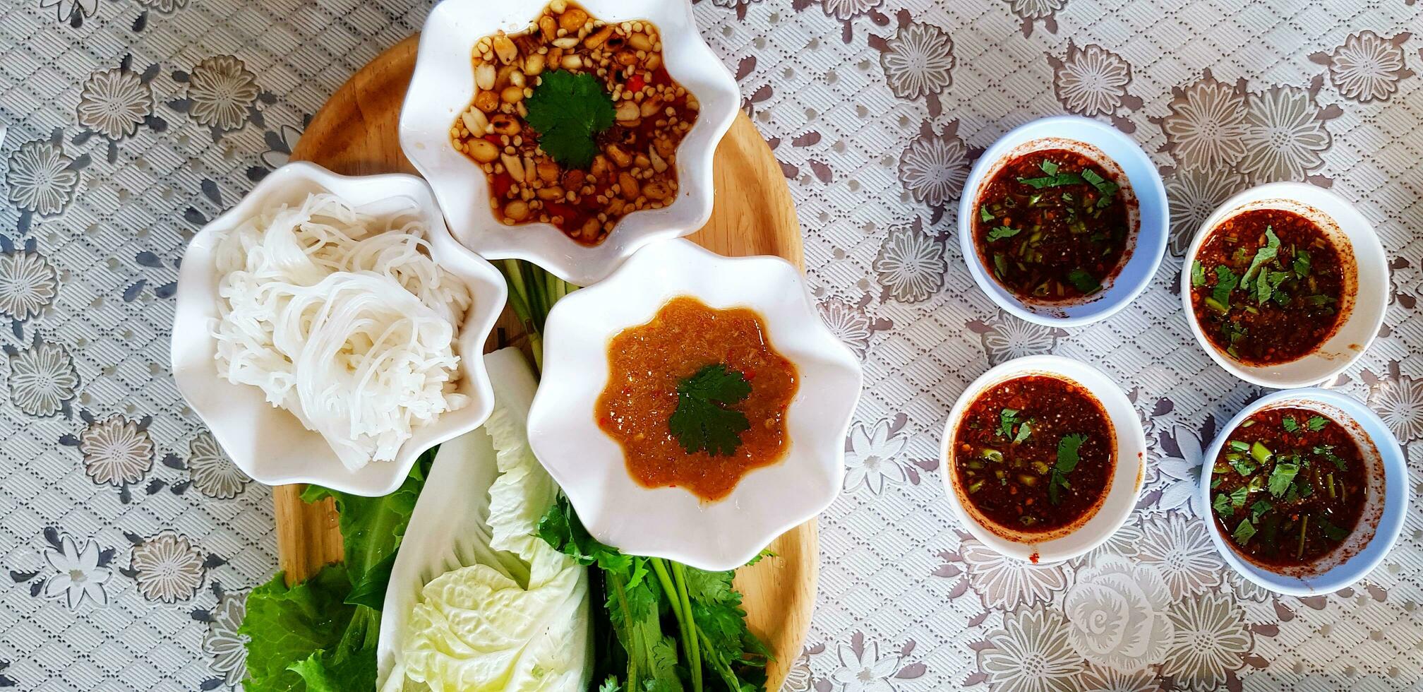 Top view of white rice noodles with fresh vegetables and sweet sauce in wooden tray with four spicy sauce on table for lunch at Thai local restaurant. Preparation food, diet lifestyle and Service photo