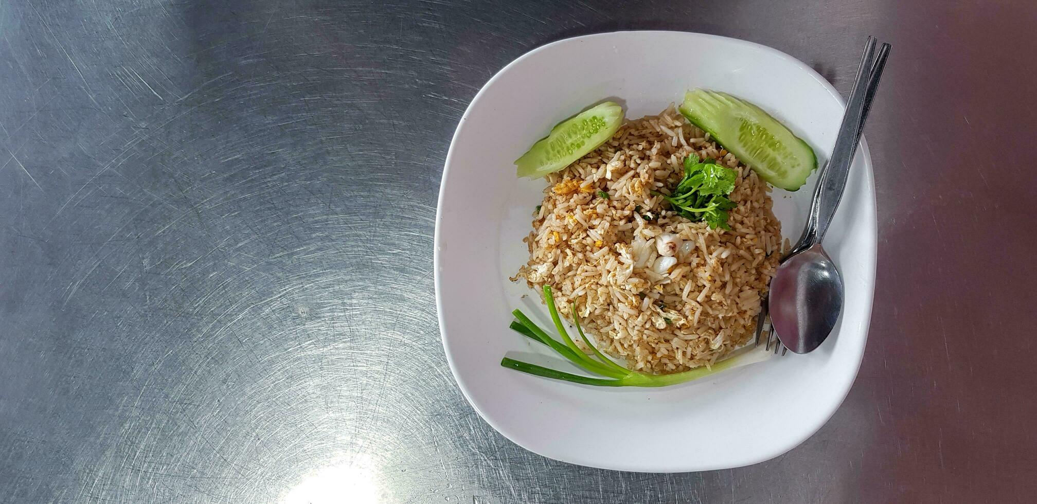 Top view or flat lay of Crab meat fried rice with slices cucumber, coriander, spring onion or scallion and spoon with fork in white dish on stainless steel table or background with left copy space photo