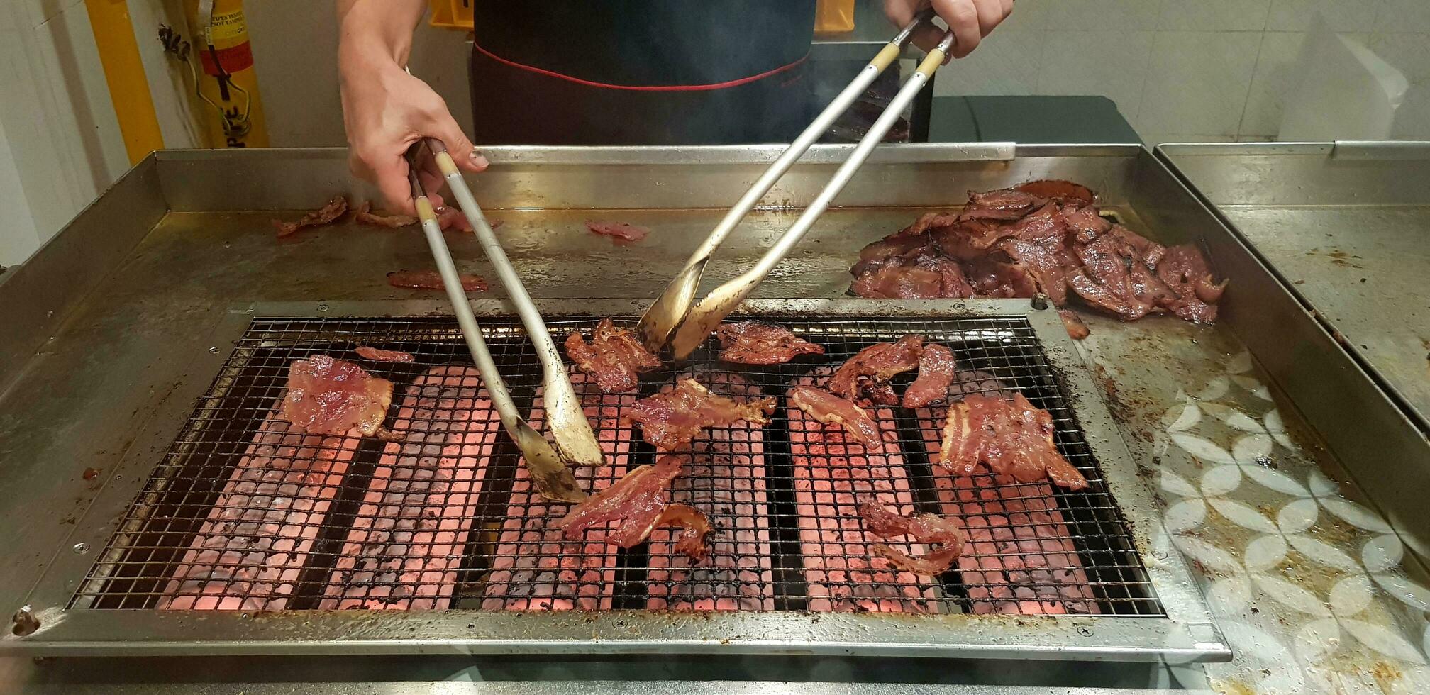 Close up many grilled red pork or barbecue food on the net stove with movement of chef's hand using utensil in the kitchen at Singapore. Selective focus and Delicious concept photo