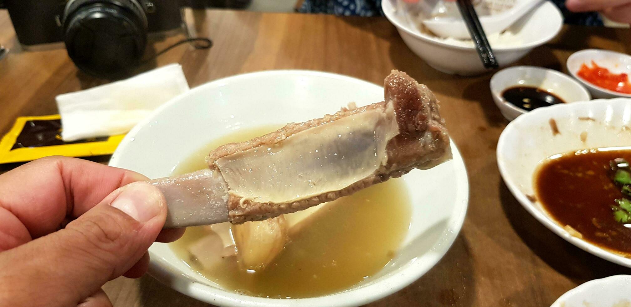 Close up hand holding boiled pork bone with hot soup bowl, sweet sauce and black camera background on wooden table at Chinese restaurant in Singapore. Asian food and Delicious dish photo
