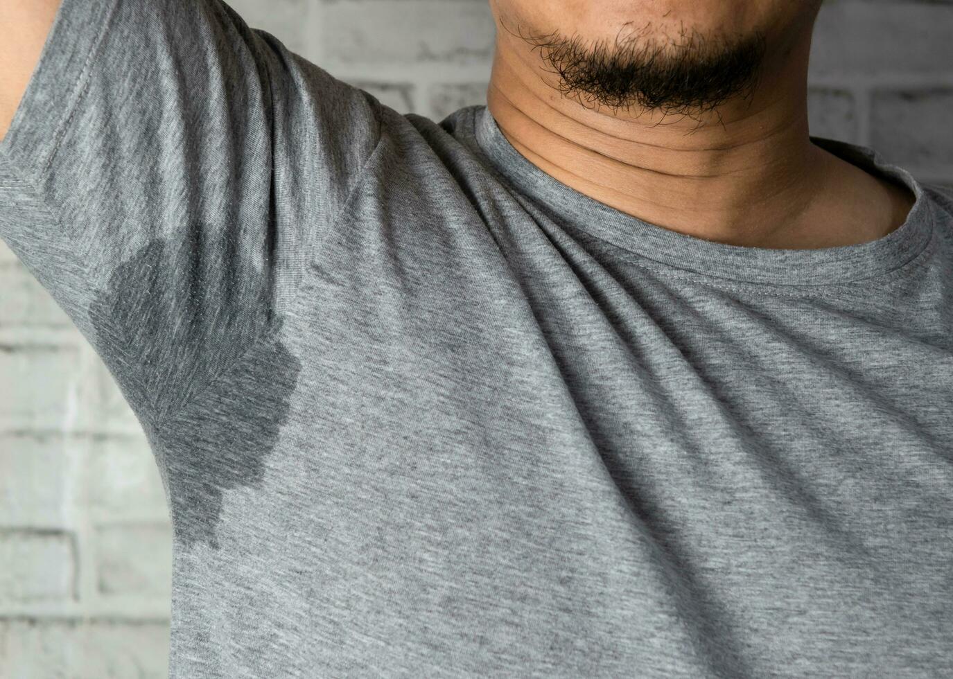 Sweaty man with stain wet armpit on t-shirt against gray photo