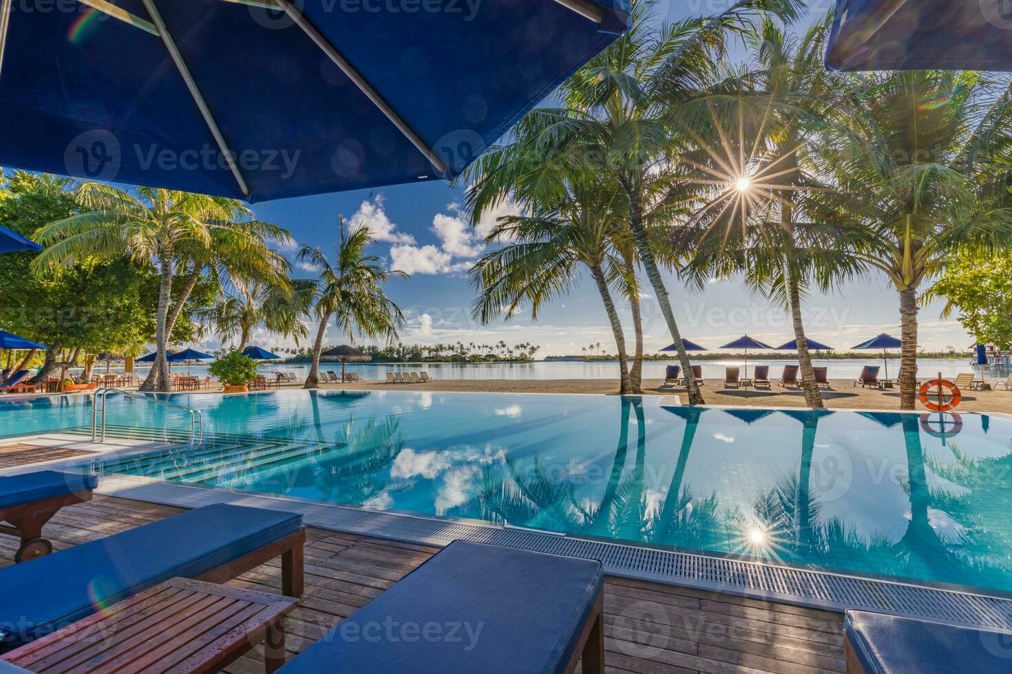 Fantastic summer mood with sun rays and palm trees over luxury infinity pool close to loungers and umbrella. Beautiful poolside and sunset sky. Luxurious tropical beach landscape. Luxury beach resort photo