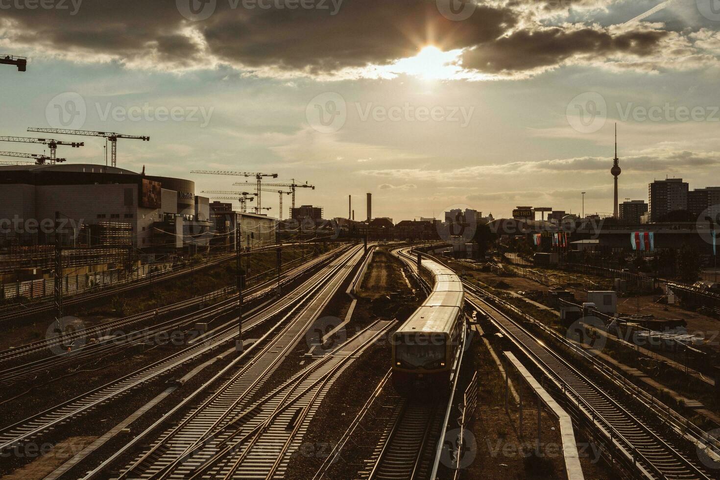 Train leaving the Berlin train station with the backlight of the golden sunset, with railyard around and the silhouette of the city and cranes in the background photo