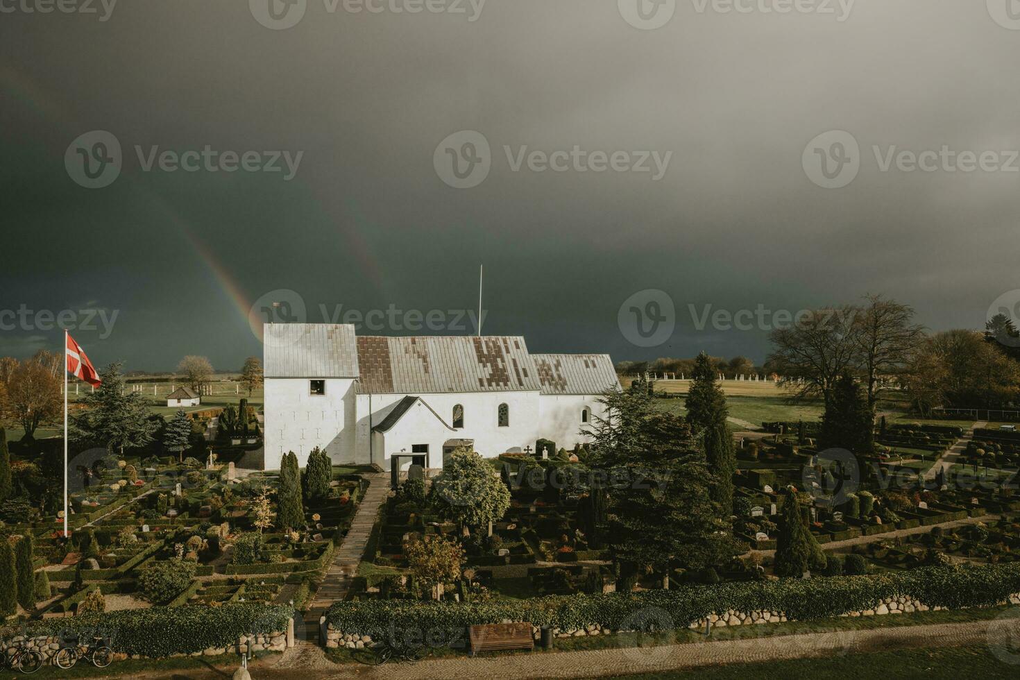 The white old church on Jelling cemetery with Denmark flag in Denmark with oldest Vikings rune stones during the dark overcast autumn day photo