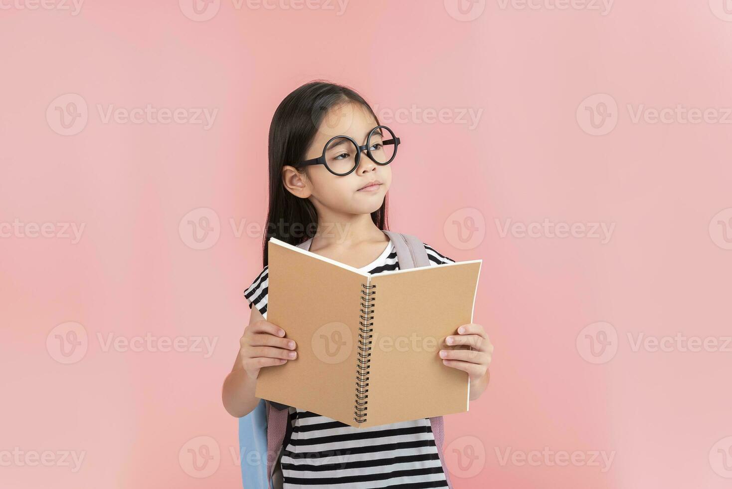 schoolgirl hugging book wearing backpack smiling isolated on pink background photo