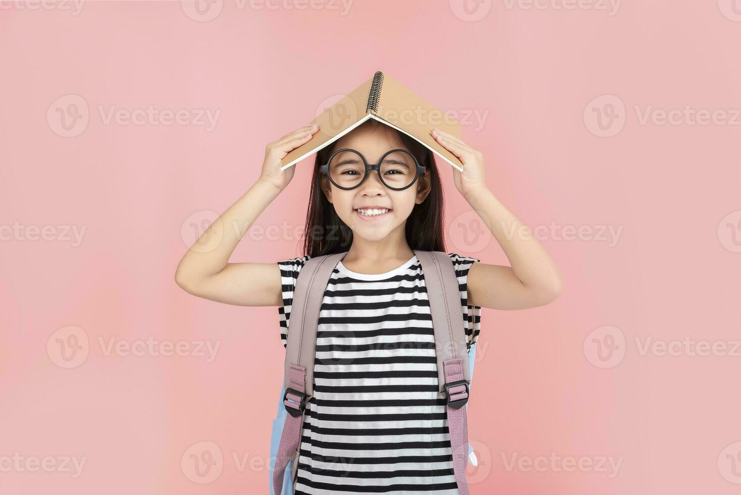 schoolgirl hugging book wearing backpack smiling isolated on pink background photo