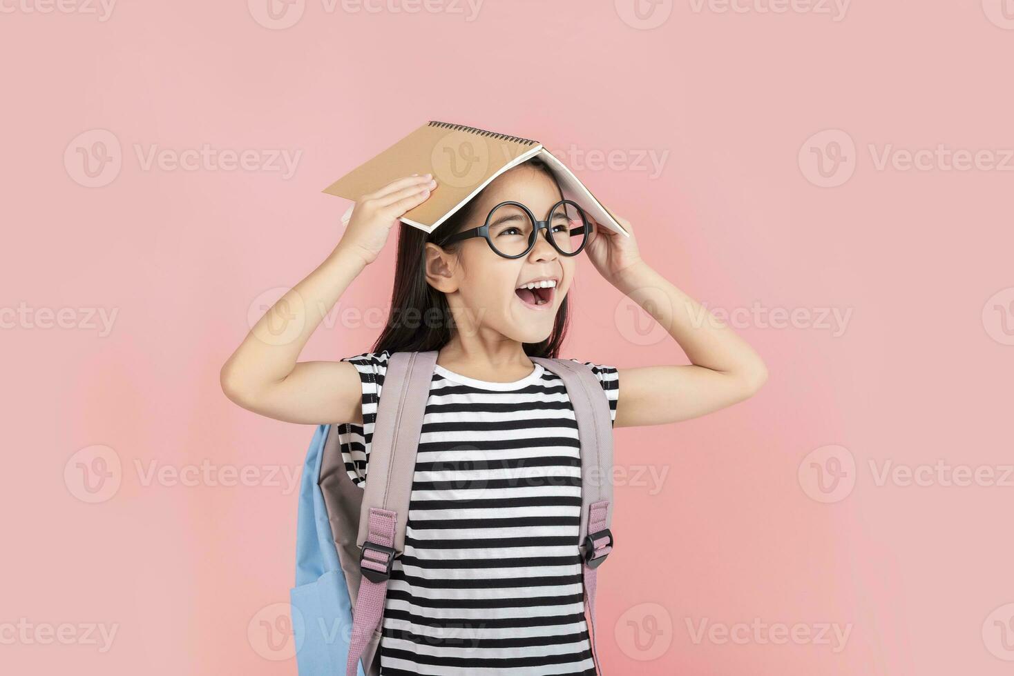 schoolgirl hugging book wearing backpack smiling isolated on pink background photo
