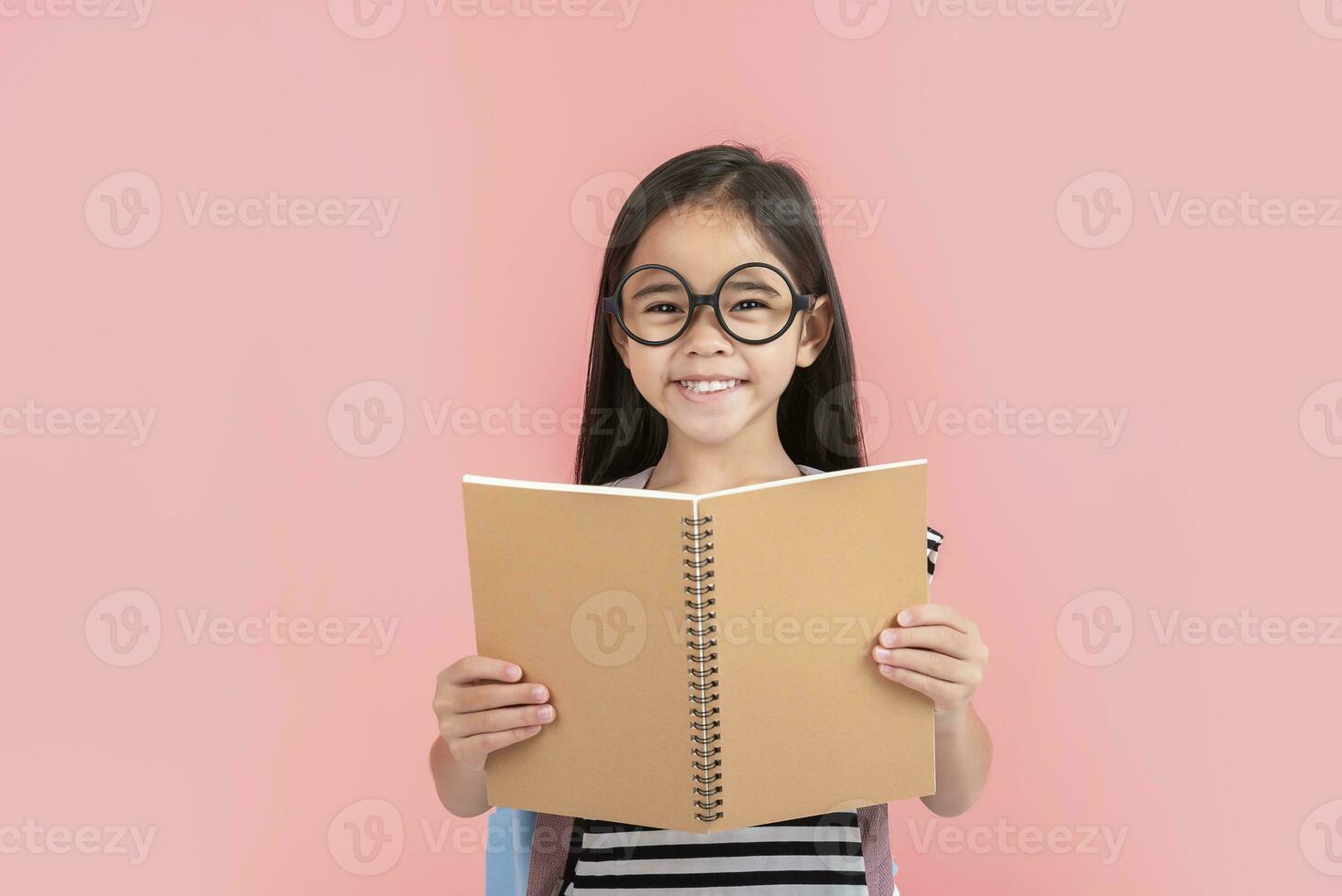 schoolgirl hugging book wearing backpack smiling isolated on pink background photo