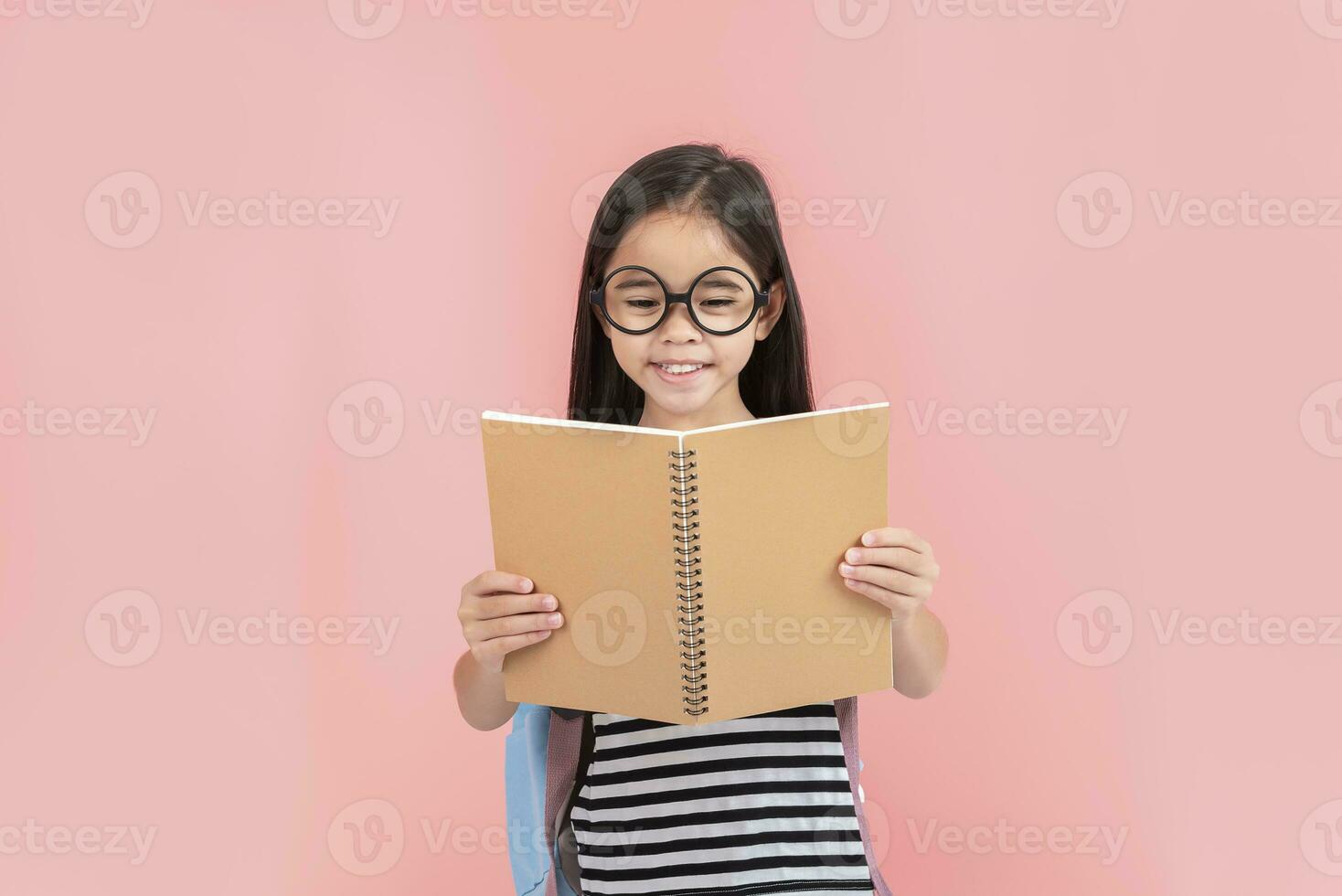 schoolgirl hugging book wearing backpack smiling isolated on pink background photo