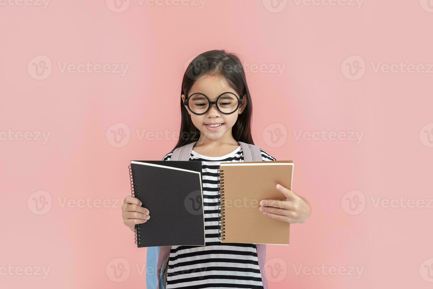 schoolgirl hugging book wearing backpack smiling isolated on pink background photo
