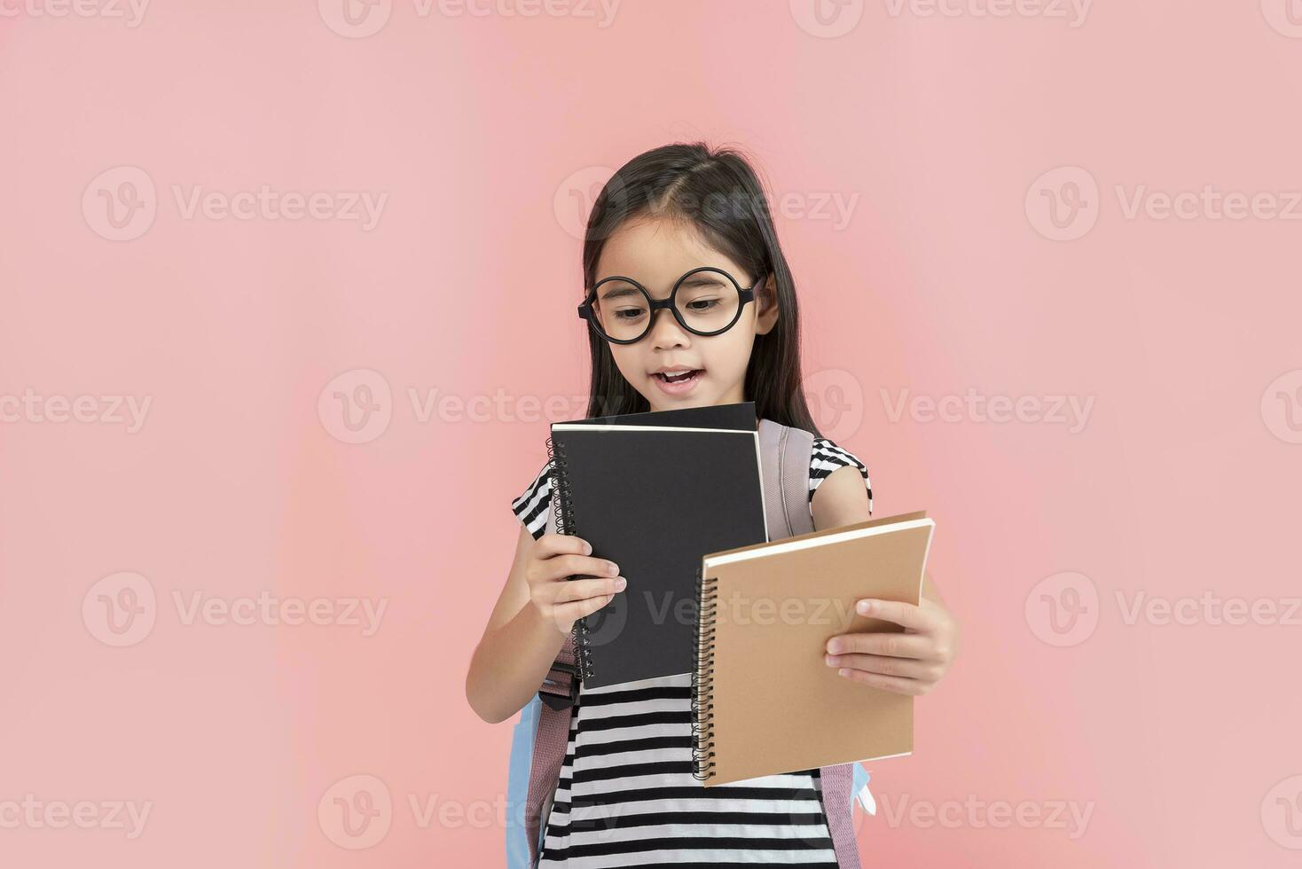 schoolgirl hugging book wearing backpack smiling isolated on pink background photo