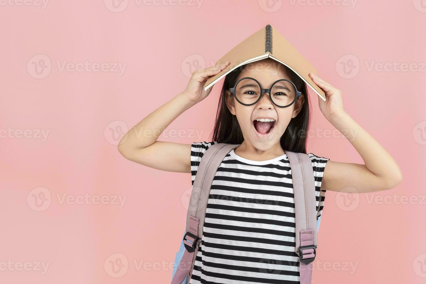 schoolgirl hugging book wearing backpack smiling isolated on pink background photo