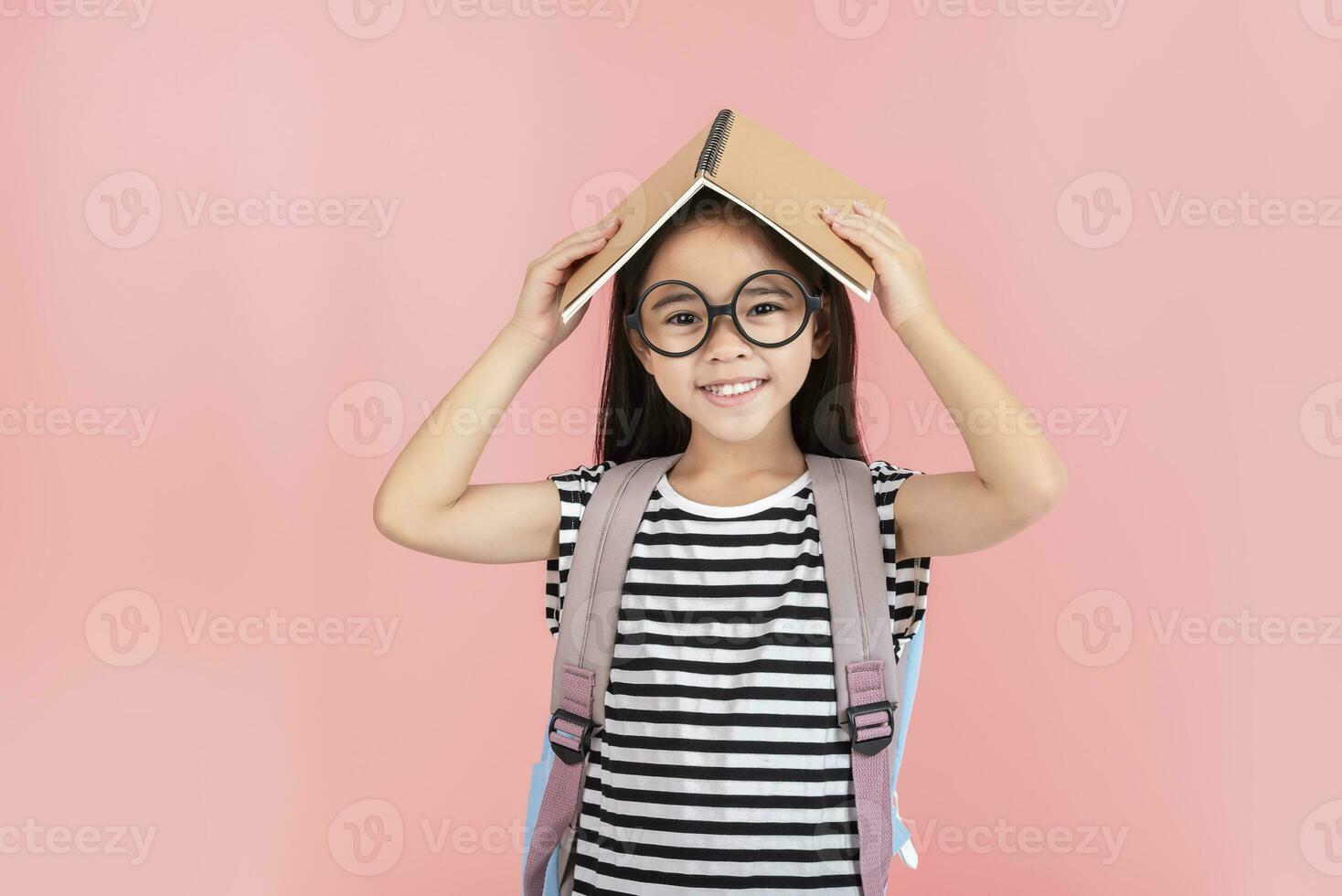 schoolgirl hugging book wearing backpack smiling isolated on pink background photo