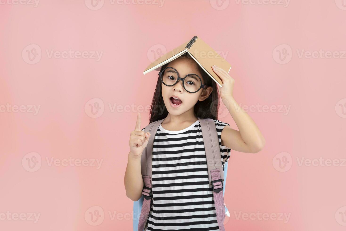 schoolgirl hugging book wearing backpack smiling isolated on pink background photo