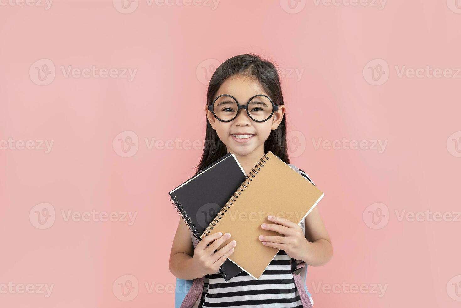 schoolgirl hugging book wearing backpack smiling isolated on pink background photo