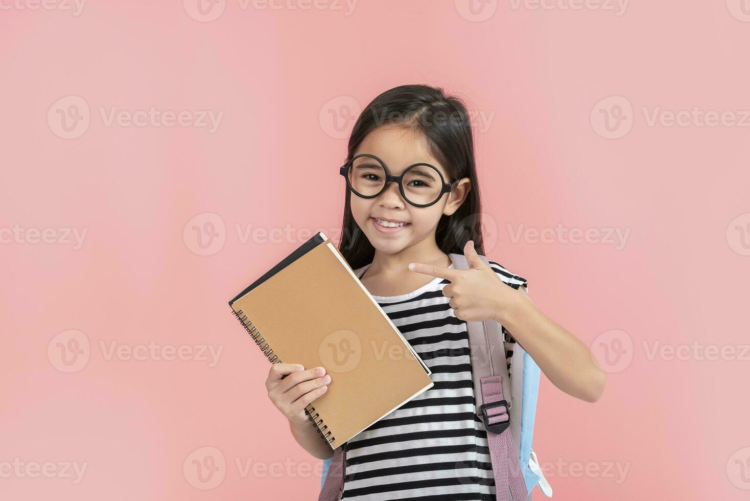 schoolgirl hugging book wearing backpack smiling isolated on pink background photo