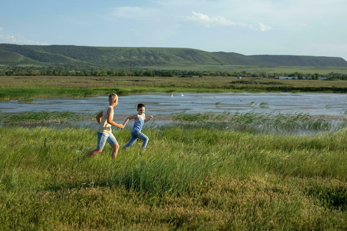 Two brothers in jeans and t-shirts run along the pond competing in speed. Summer sunny day near the pond. Summer holidays in the countryside photo