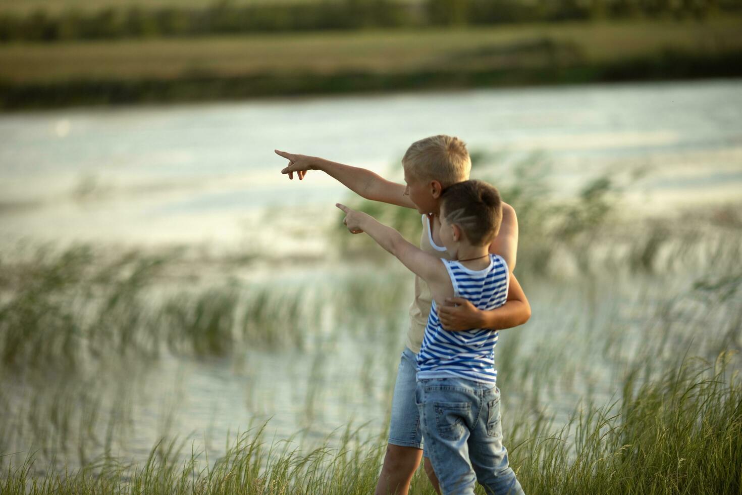 Two boys in jeans and t-shirts point a finger into the distance near the pond. Summer sunny day near the pond. Summer holidays in the countryside. Happy childhood photo