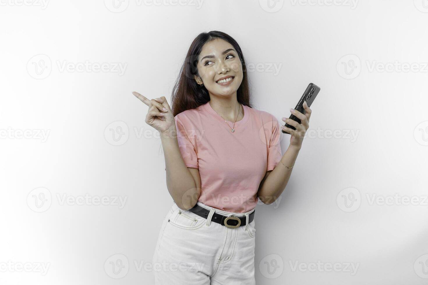 A portrait of a happy Asian woman wearing pink t-shirt, holding her phone, and pointing copy space on beside her, isolated by white background photo