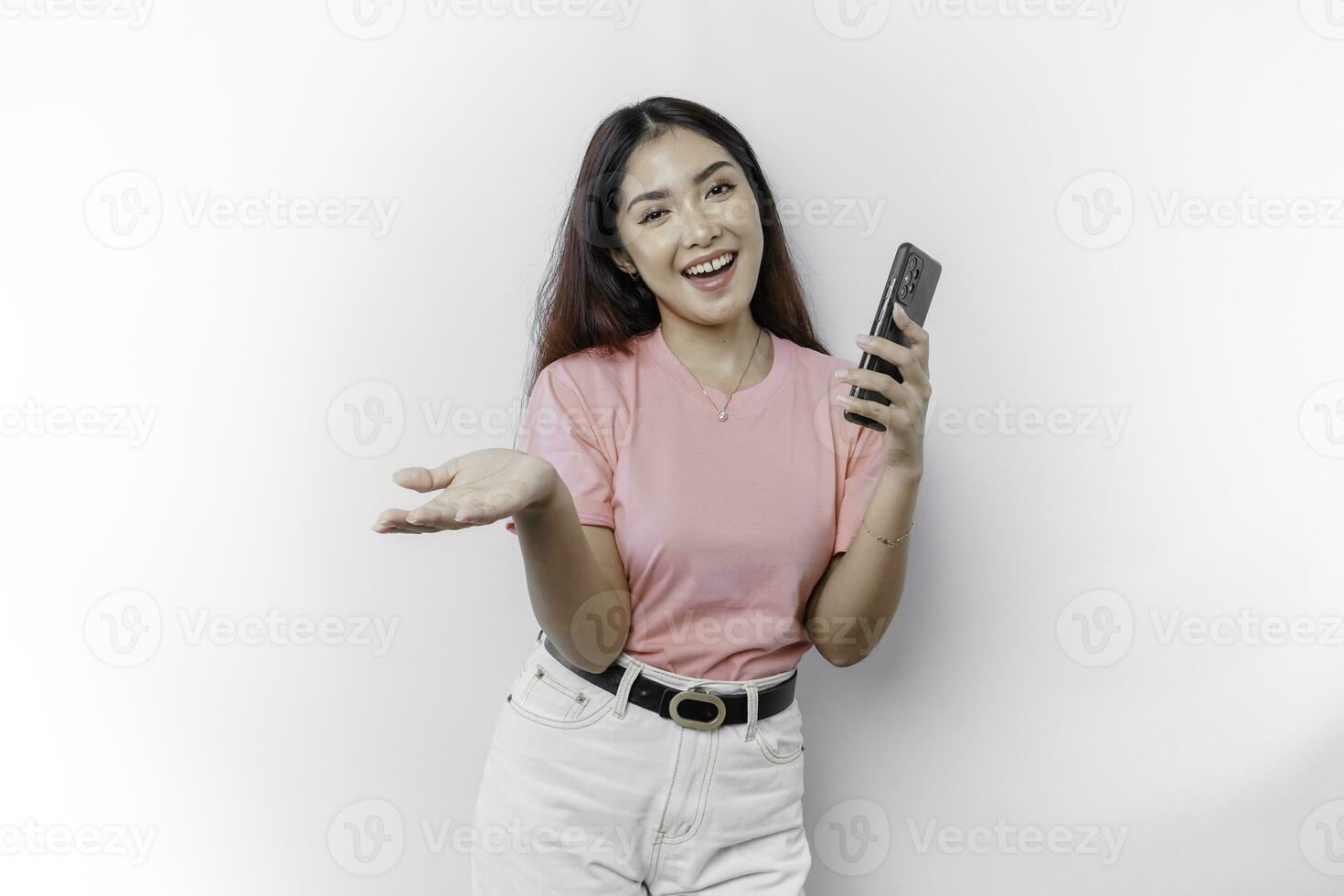 A portrait of a happy Asian woman wearing pink t-shirt, holding her phone, and pointing copy space on beside her, isolated by white background photo