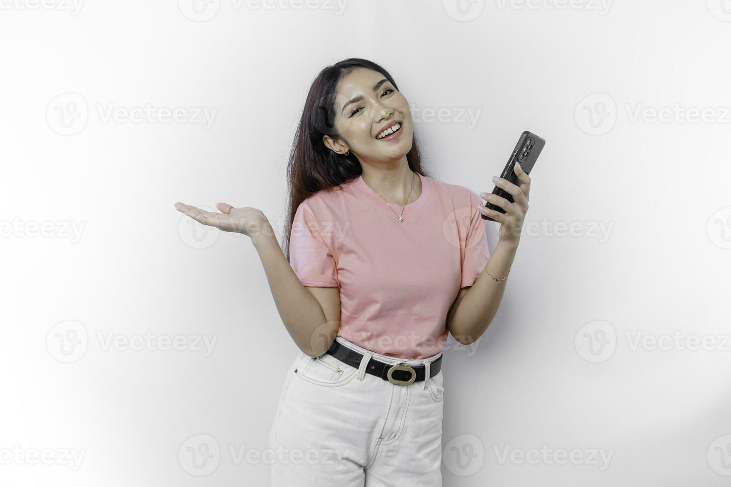 A portrait of a happy Asian woman wearing pink t-shirt, holding her phone, and pointing copy space on beside her, isolated by white background photo