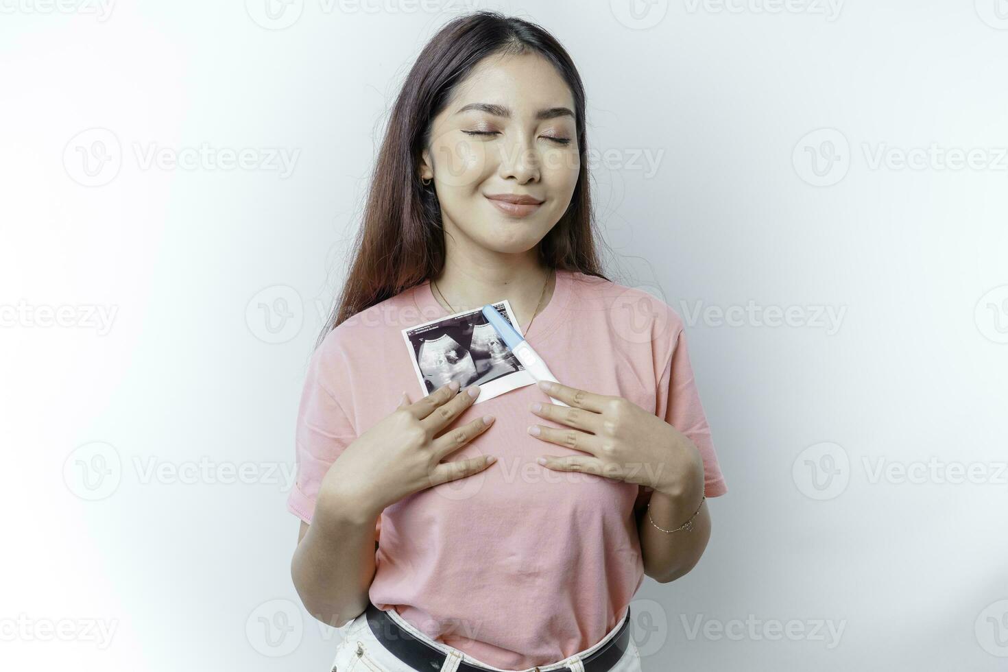 Happy young woman wearing pink t-shirt showing her pregnancy test and ultrasound picture, isolated on white background, pregnancy concept photo