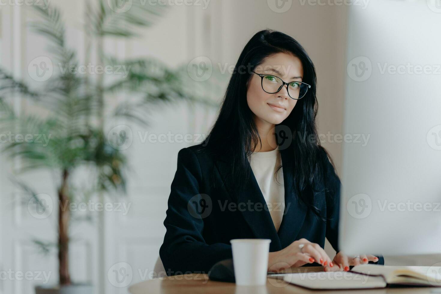 Serious successful female office worker in formal outfit, wears eyeglasses, busy with online work, poses at desktop, works on computer, reads business new or watches tutorial, enjoys working process. photo