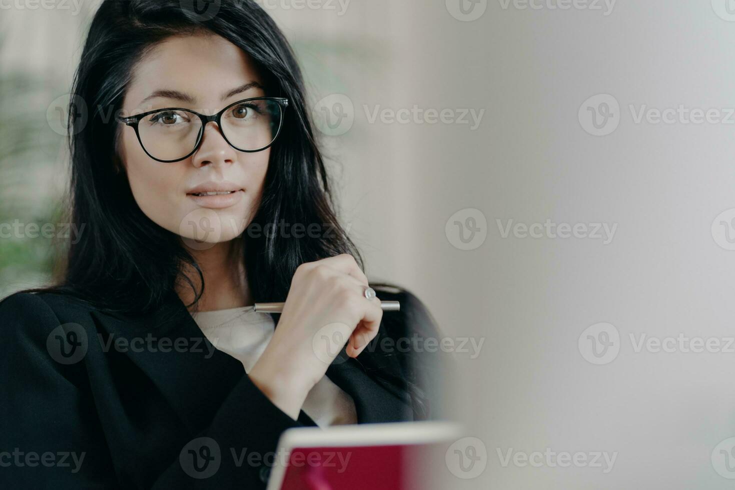 Self confident serious adult female employee looks though transparent glasses, writes information while working on project, makes plans, dressed in formal wear, notes some records in notepad photo