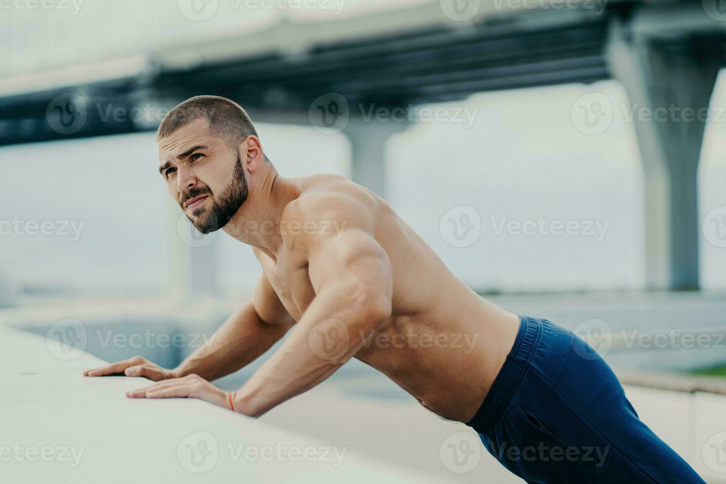 Bearded man does push-ups outdoors, warms up, poses near a bridge, has early morning workout. photo