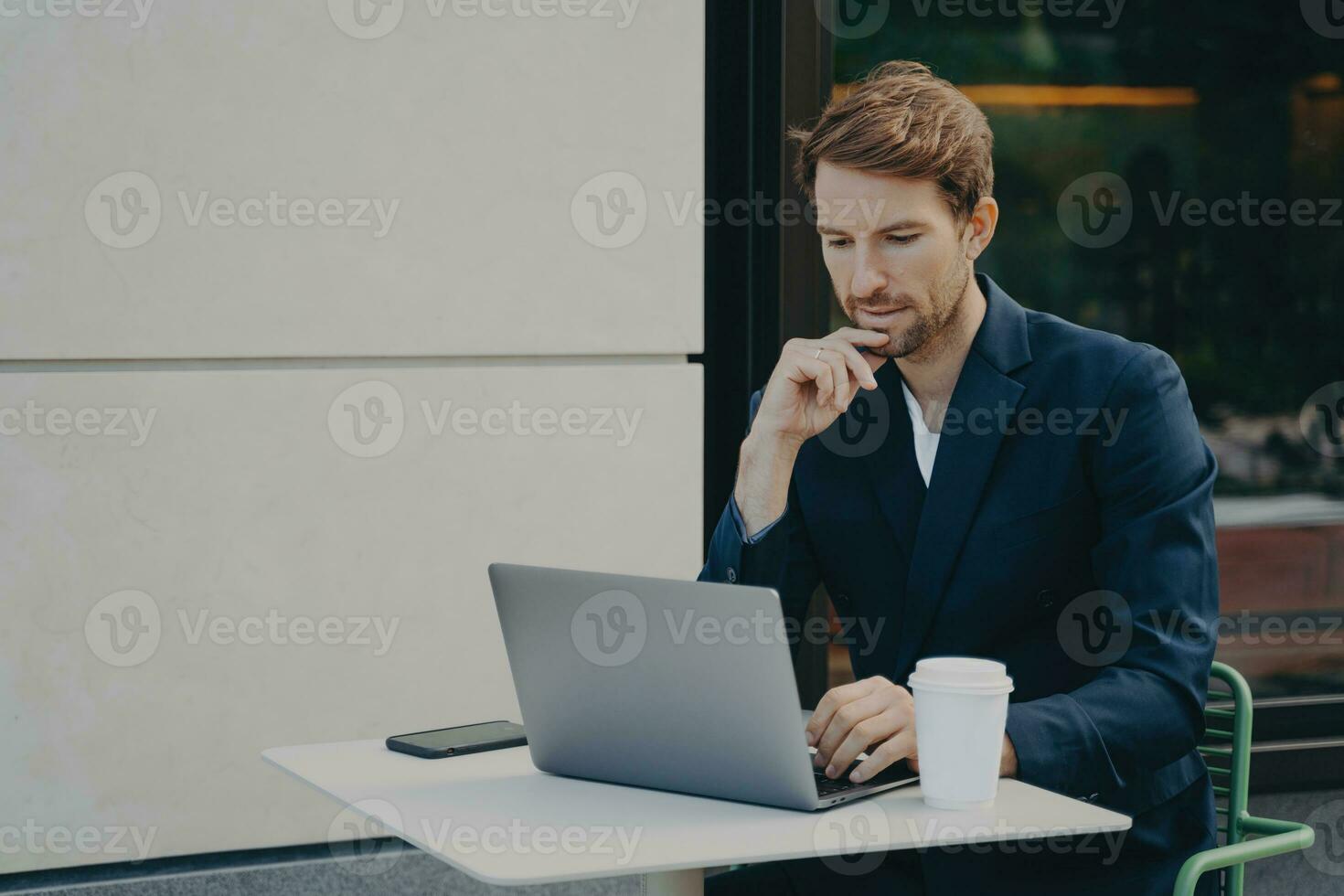 Handsome pensive young man browsing web on laptop while working remotely in sidewalk cafe photo