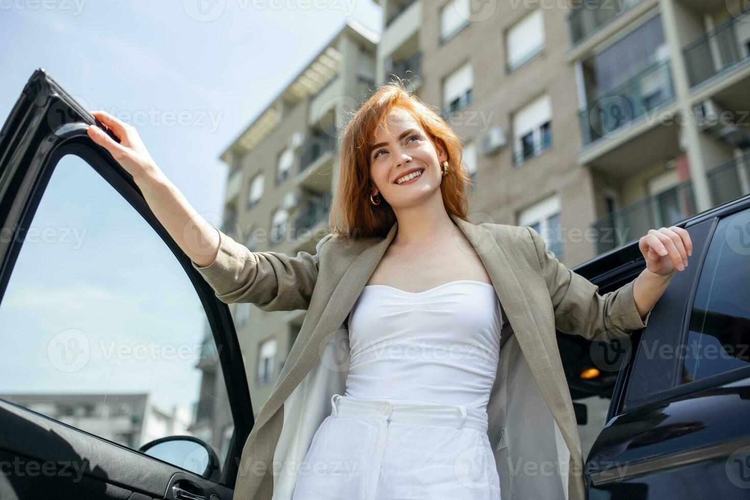 Stunning young woman in entering her car holding the door with one hand and leaning on the hood with the other photo