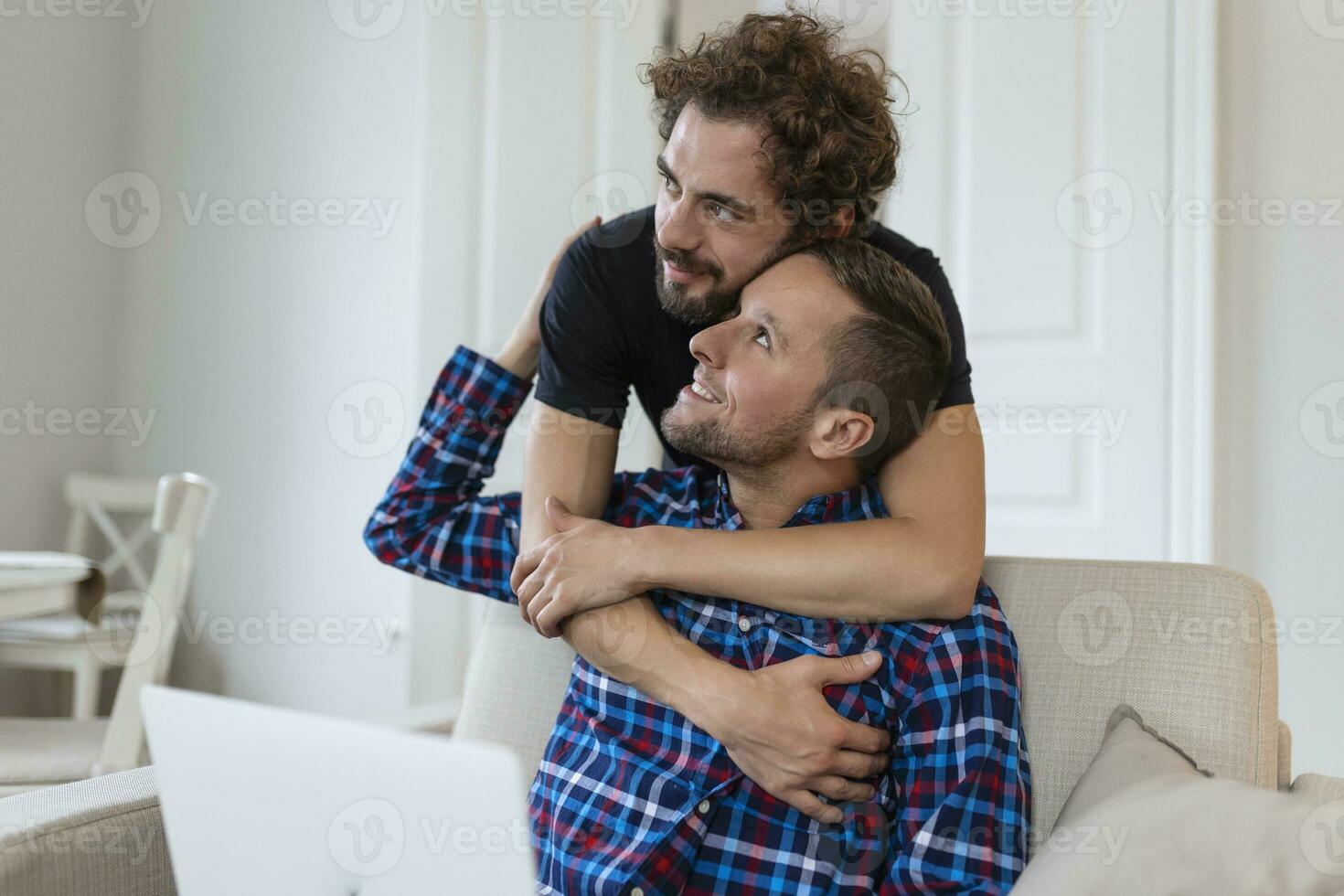 encantador gay Pareja riendo juntos mientras sentado en su vivo habitación a hogar. dos romántico joven masculino amantes teniendo divertido surf el Internet adentro. joven gay Pareja vivo juntos. foto