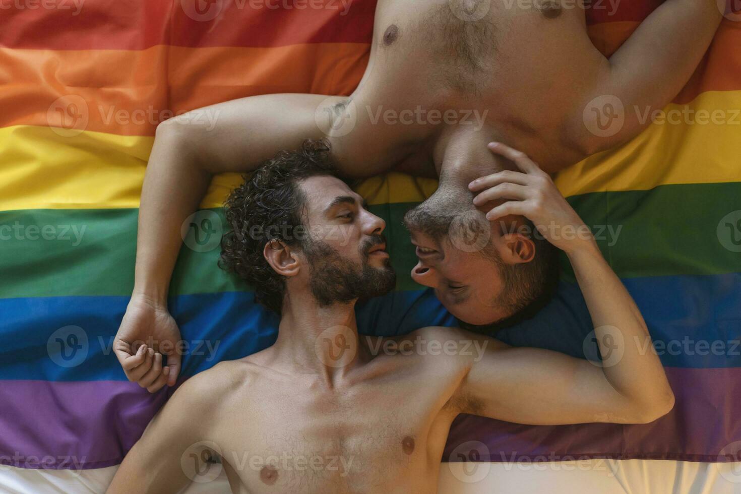 cariñoso joven gay hombre besos su amante en el cama. dos joven masculino amantes tendido juntos en orgullo bandera. romántico joven gay Pareja unión afectuosamente adentro. foto