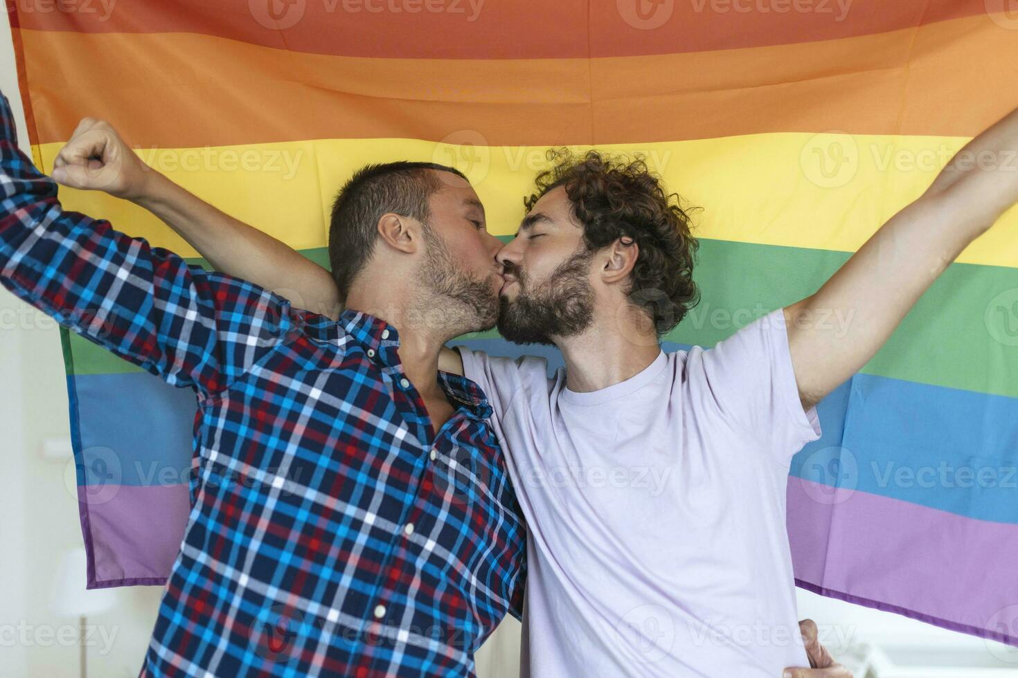 dos joven gay amantes besos cada otro afectuosamente. dos joven masculino amantes en pie juntos en contra un orgullo bandera. cariñoso joven gay Pareja compartiendo un romántico momento juntos. foto