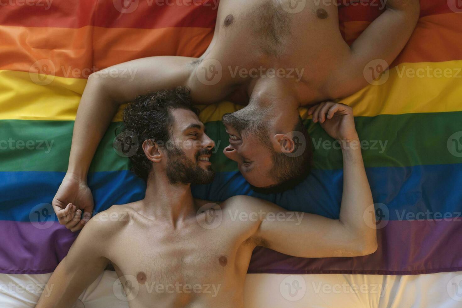 Affectionate young gay man kissing his lover on the bed. Two young male lovers laying together on pride flag. Romantic young gay couple bonding fondly indoors. photo