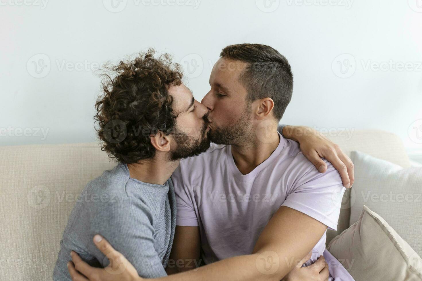 encantador gay Pareja besos mientras sentado en su vivo habitación a hogar. dos romántico joven masculino amantes teniendo divertido adentro. joven gay Pareja vivo juntos. foto