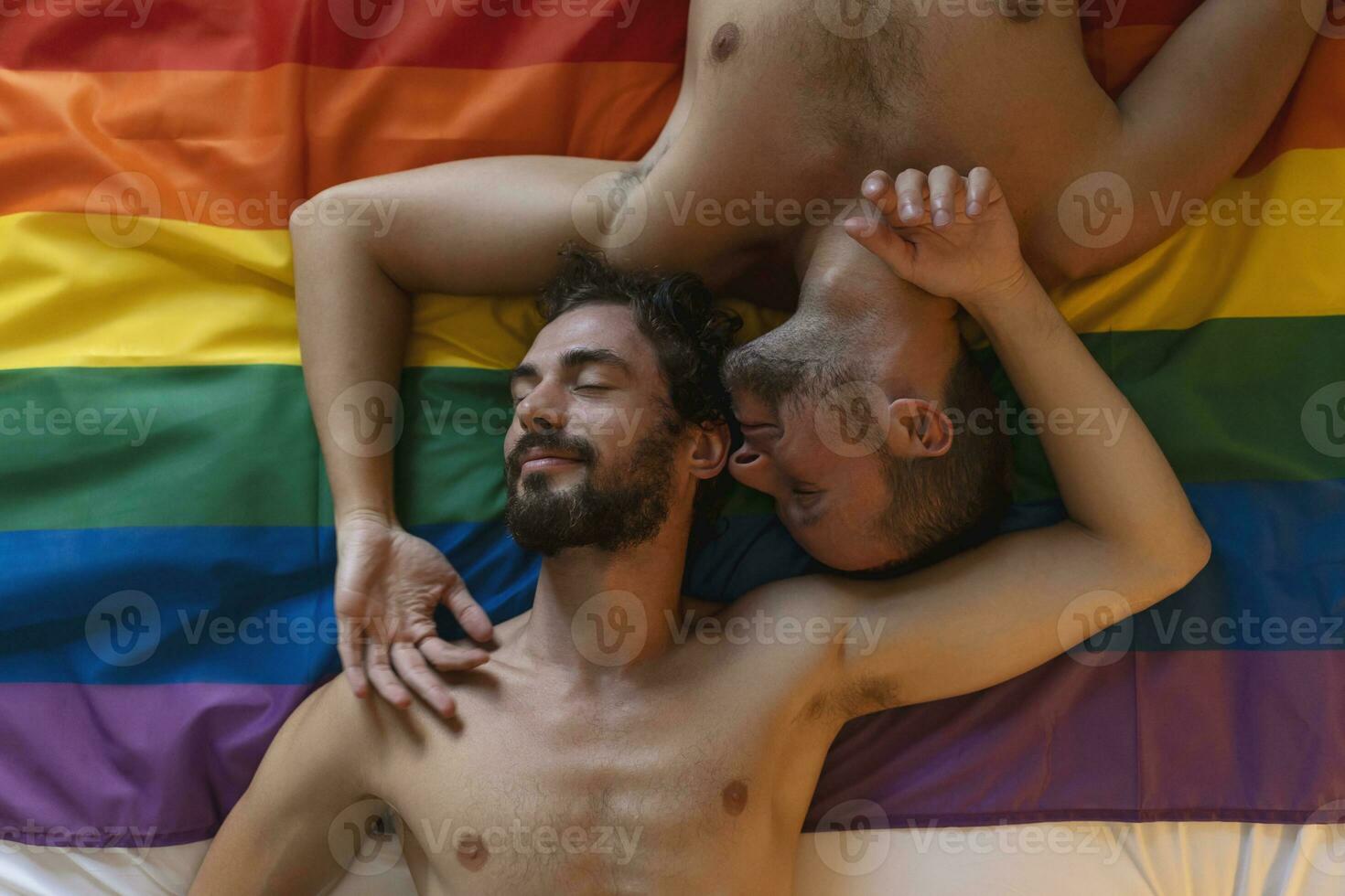 cariñoso joven gay hombre besos su amante en el cama. dos joven masculino amantes tendido juntos en orgullo bandera. romántico joven gay Pareja unión afectuosamente adentro. foto