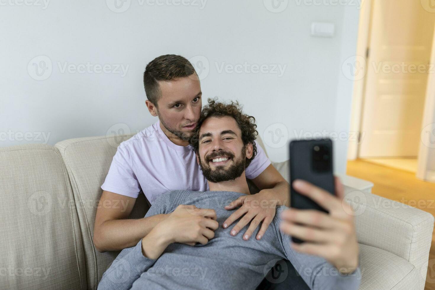 linda joven gay Pareja vídeo vocación su amigos en su vivo habitación a hogar. dos masculino amantes sonriente alegremente mientras saludo su amigos en un teléfono inteligente joven gay Pareja sentado juntos. foto