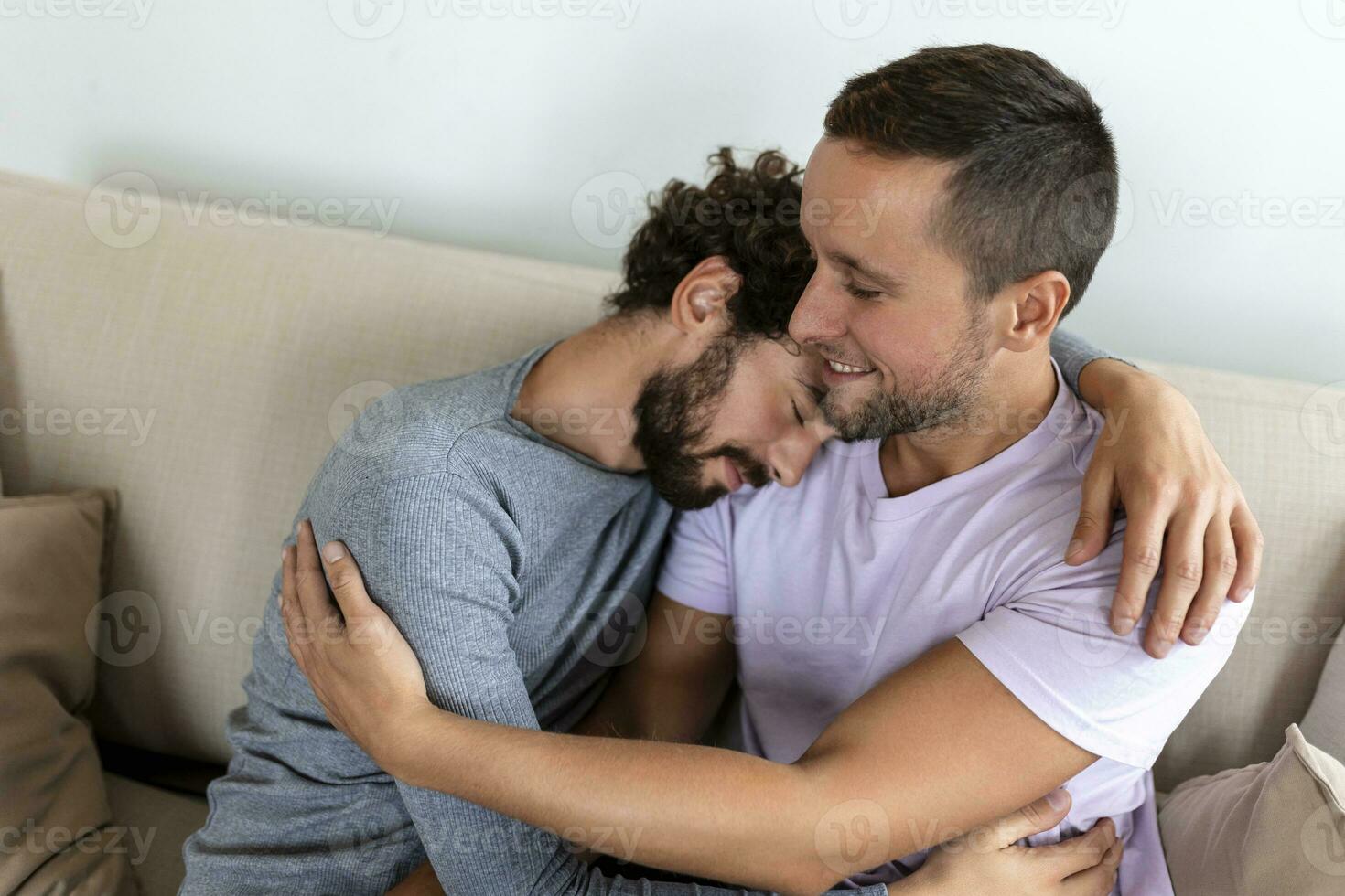 encantador gay Pareja besos mientras sentado en su vivo habitación a hogar. dos romántico joven masculino amantes teniendo divertido adentro. joven gay Pareja vivo juntos. foto