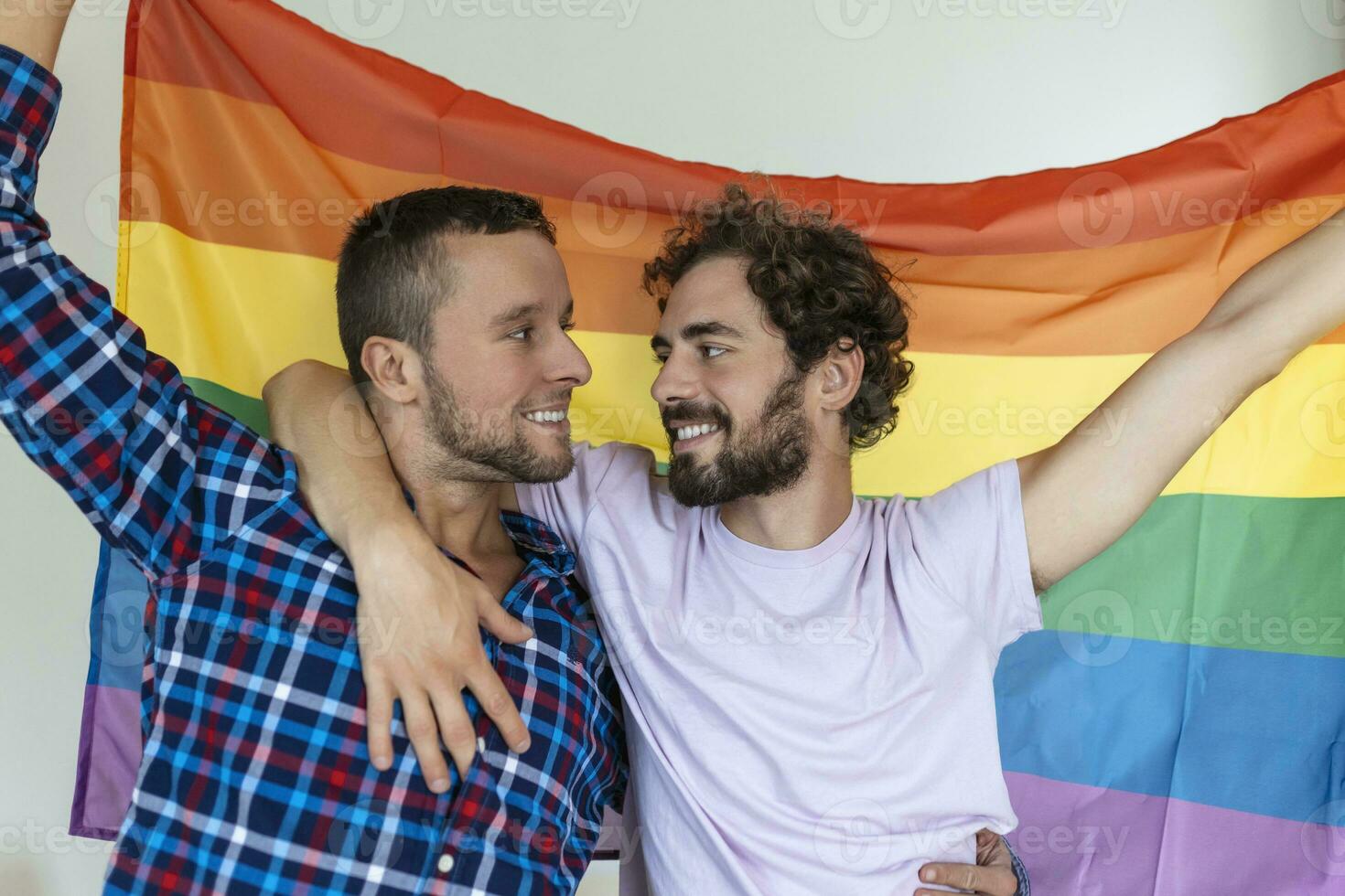 Two young gay lovers looking at each other affectionately. Two young male lovers standing together against a pride flag. Affectionate young gay couple sharing a romantic moment together. photo