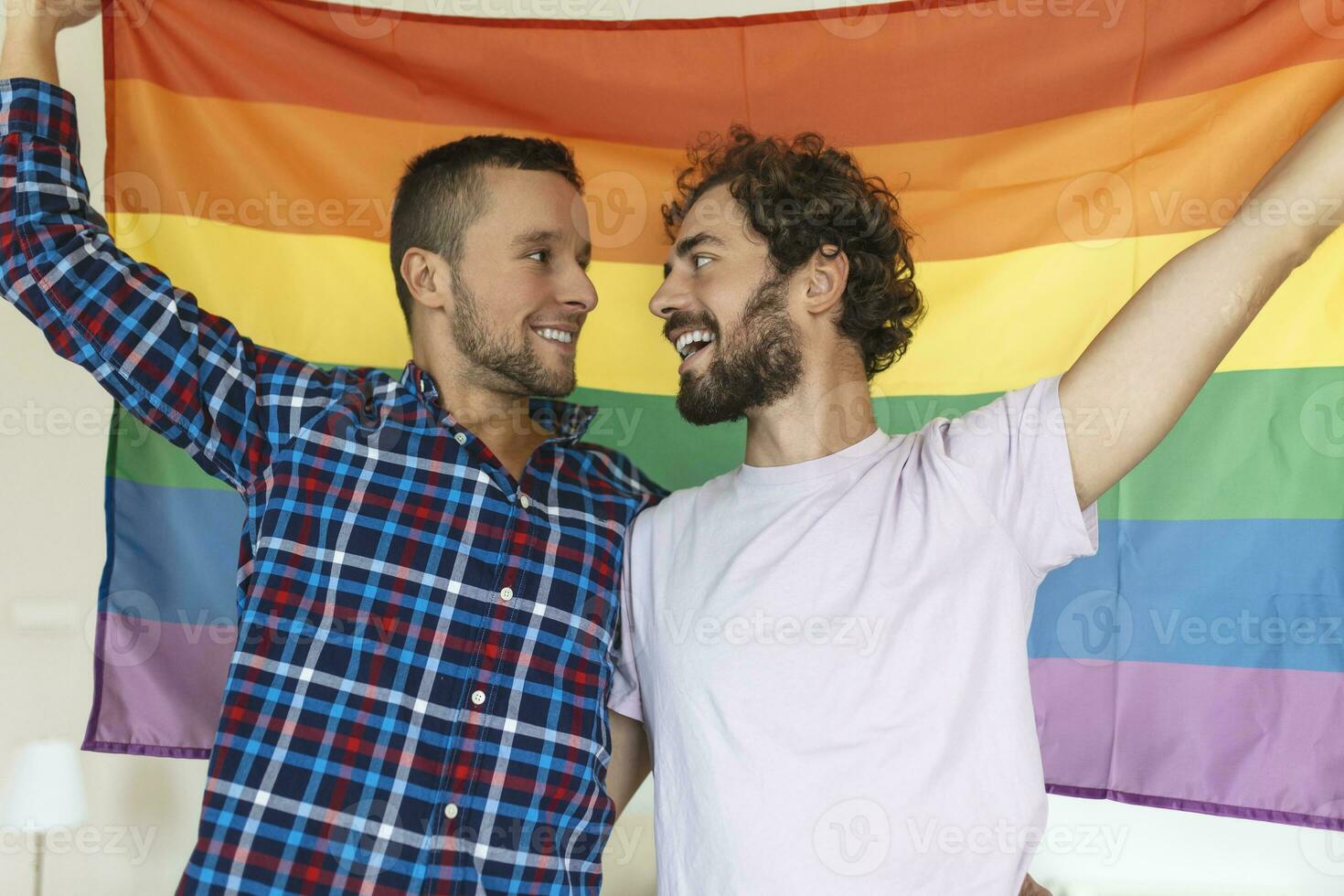 dos joven gay amantes mirando a cada otro afectuosamente. dos joven masculino amantes en pie juntos en contra un orgullo bandera. cariñoso joven gay Pareja compartiendo un romántico momento juntos. foto