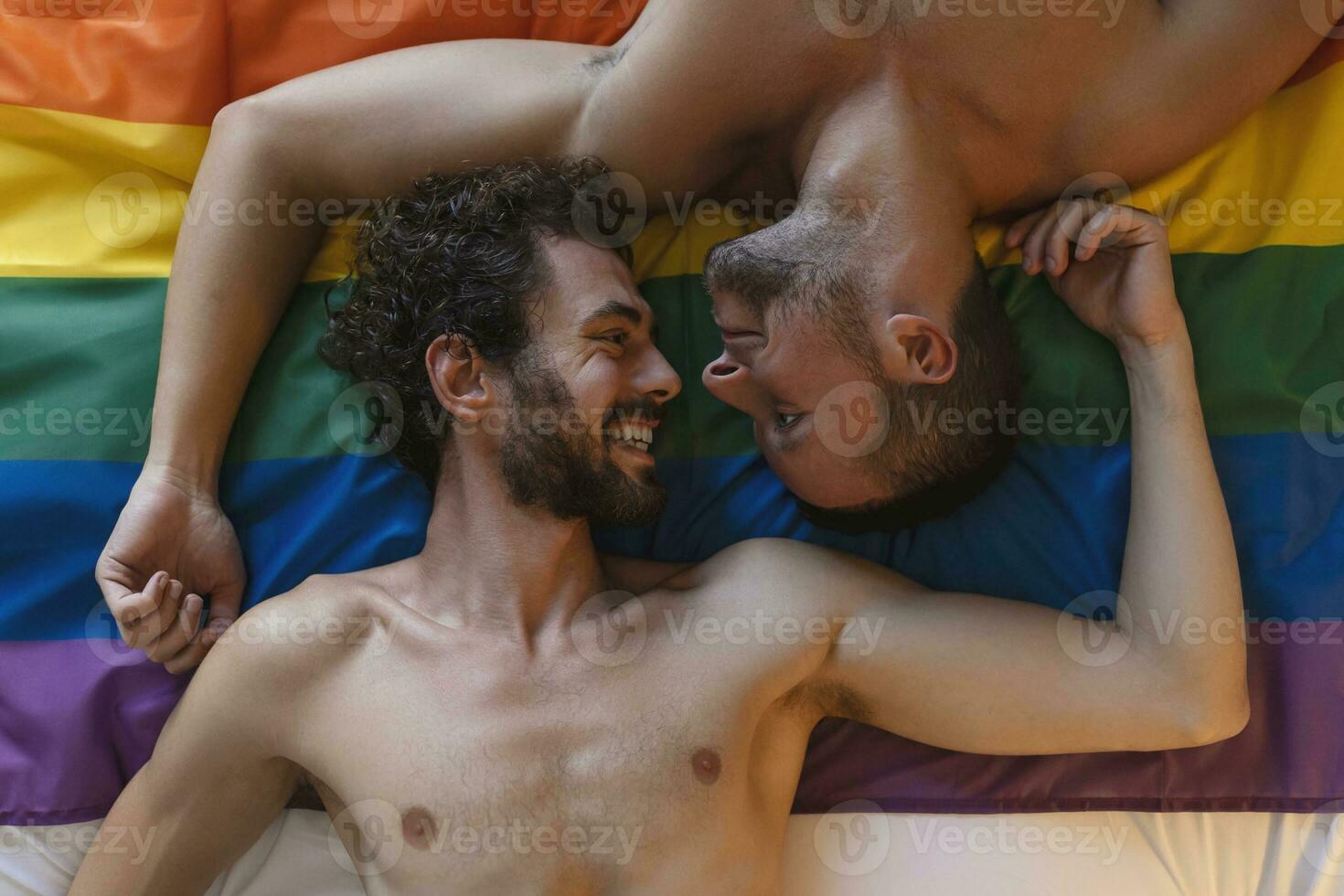 cariñoso joven gay hombre besos su amante en el cama. dos joven masculino amantes tendido juntos en orgullo bandera. romántico joven gay Pareja unión afectuosamente adentro. foto