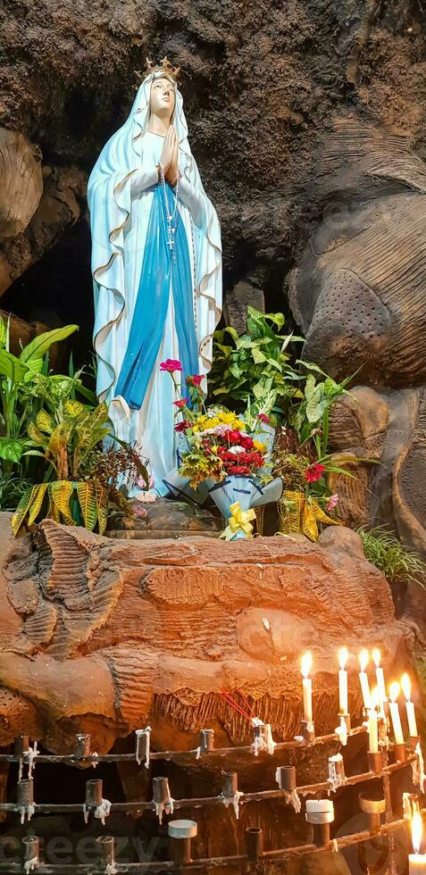 Statue of Holy Virgin Mary in Roman Catholic Church, in the cave of virgin mary, in a rock cave chapel Catholic Church with tropical flowers around photo
