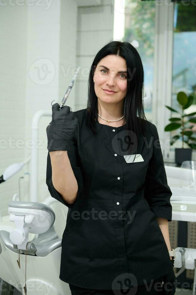 Confident female dental hygienist holding a syringe with dental anesthesia, looking at camera, standing by dentist chair photo