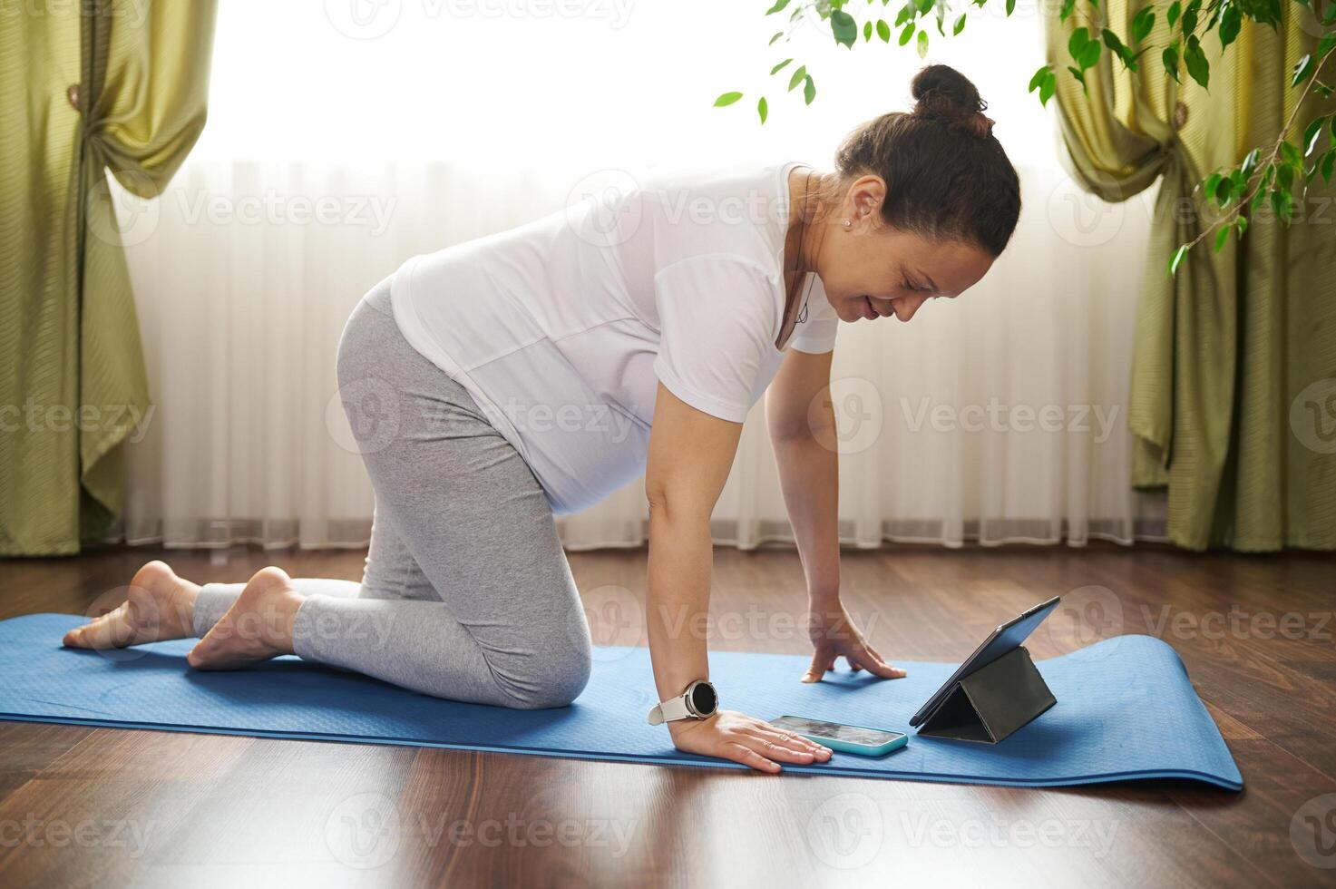 Pregnant woman using mobile phone and tablet during online yoga practice, standing in table posture on blue exercise mat photo