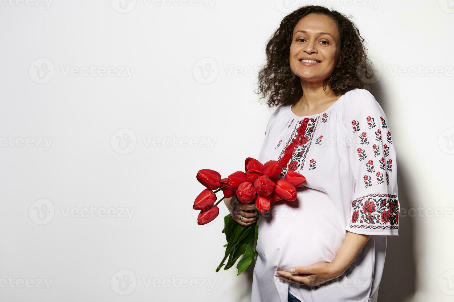 encantador adulto embarazada mujer en ucranio étnico bordado camisa, sonriente, acariciando barriga, posando con rojo tulipanes foto