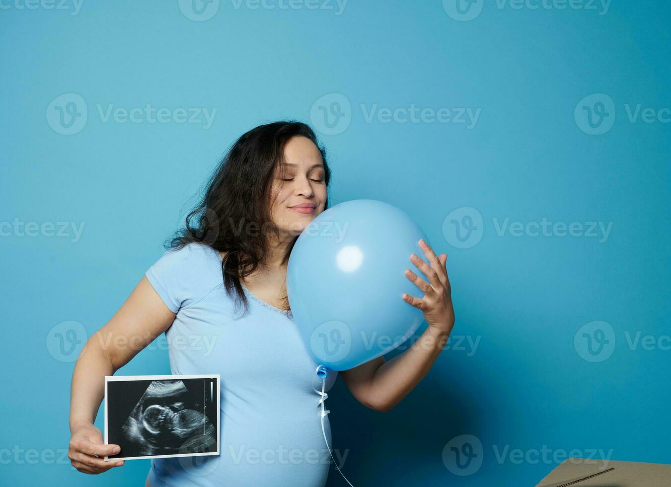 encantador embarazada mujer abrazando azul globo, posando con ultrasonografía imagen de su futuro bebé chico, azul fondo foto