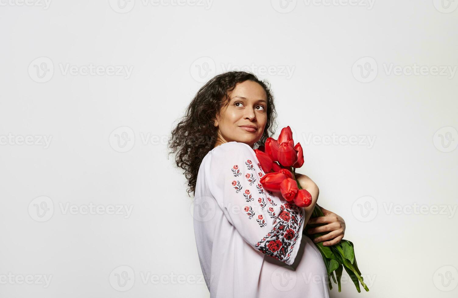 Happy pregnant woman in embroidered shirt with ukrainian ethnic ornament, holding bunch of red tulips, isolated on white photo