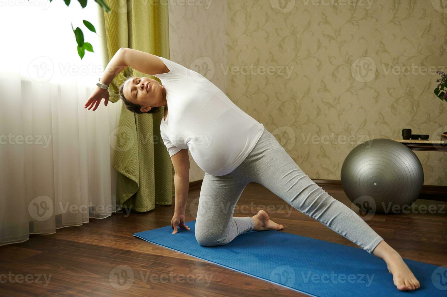 Active pregnant woman with big belly, stretching her body while practicing prenatal fitness on blue exercise mat at home photo