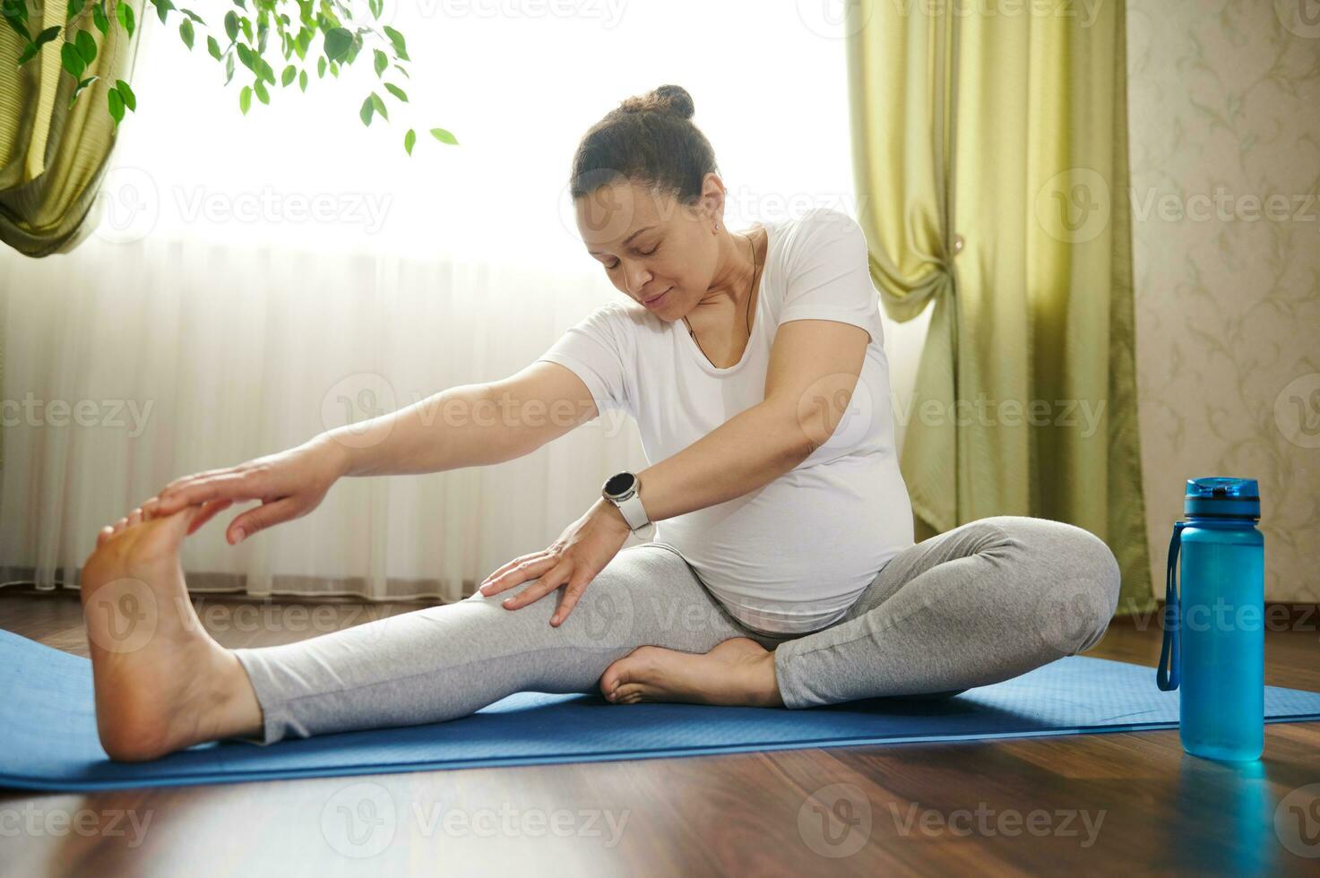 embarazada mujer alcanzando brazo a su pierna, haciendo prenatal extensión ejercicios en aptitud estera, practicando yoga a hogar. foto